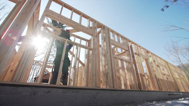 Sliding view of builder working at frame residential house building, attaching wooden planks with hydraulic hammer. Sunny winter day