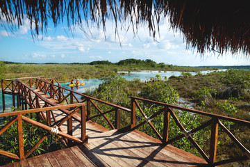 Fototapeta na wymiar Jetty along seven colored lagoon with canoe passing by surrounded by tropical plants in Bacalar, Quintana Roo, Mexico