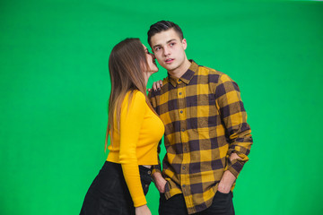 Young women sharing gossip and whispering secrets to her boyfriend, he is looking with puzzled face expression, standing over green background.