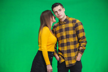 Young women sharing gossip and whispering secrets to her boyfriend, he is looking with puzzled face expression, standing over green background.