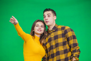 Portrait of young woman pointing at something ahead isolated on green background, her boyfriend is looking at it, pleasantly surprised.