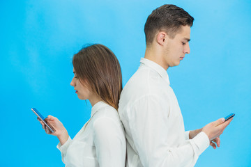 Beautiful young business woman and handsome businessman in formal clothes are using gadgets, standing back to back over blue background.