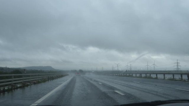 Car windshield view driving M56 highway in poor visibility conditions because of rain and spray; passing group of wind turbines. Two clips joined.