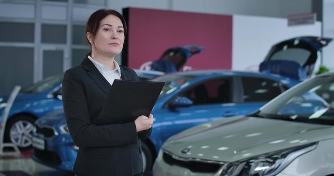 Portrait of Caucasian female car dealer standing in dealership with document holder and thinking. Serious businesswoman working in showroom. Auto industry, trading. Cinema 4k ProRes HQ.