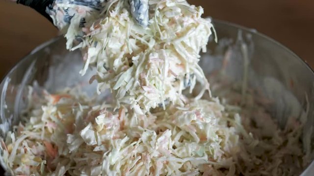 Mixing creamy homemade dressing with crips cabbage and chopped carrots into delicious coleslaw salad in a chrome bowl wearing a black glove