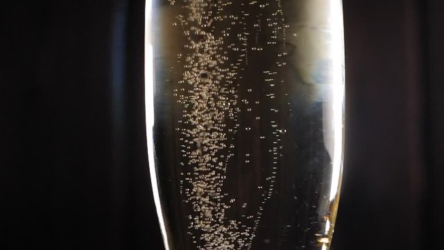 Close-up of a glass of champagne with small beautiful bubbles slowly spinning on a black background. Slow motion. Glare from the light on a glass of champagne.