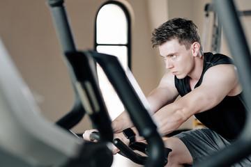 Fototapeta na wymiar Concentrated serious fit young man riding bicycle simulator in gym