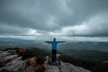 Obraz premium Adventurer on the edge of a cliff watching a wooded landscape, on a rocky mountain, rain clouds are seen in the background