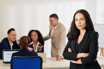 Obraz premium Beautiful successful female entrepreneur standing with her arms folded, her team discussing work in background