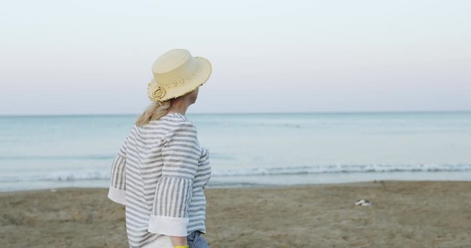 The cheerful elderly woman in straw hat walking on the sea beach in the early morning at sunrise.
