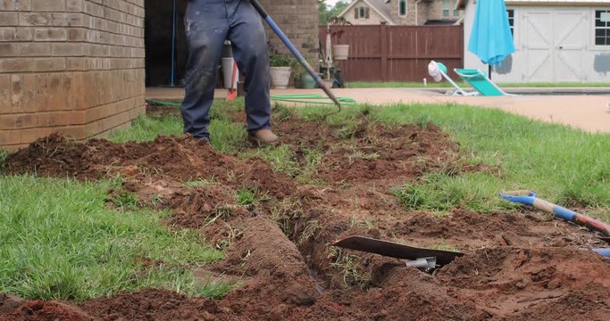 Worker rakes dirt to fill in a trench and bury a new french drain pipe system underground in a yard