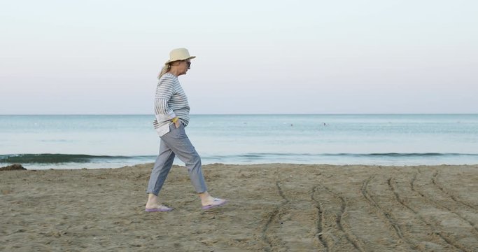 The cheerful elderly woman in straw hat walking on the sea beach in the early morning at sunrise.