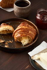 Morning breakfast with croissant on plate, cup of coffee, jam and butter. Dark brown table