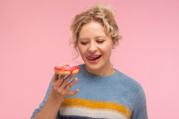 Tasty dessert. Portrait of woman with short curly hair in sweater holding doughnut, looking with desire at donut and licking her lips, wants to eat sweet confection. indoor studio shot pink background