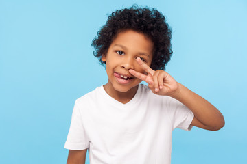 Portrait of cheerful carefree little boy with curly hair in T-shirt picking nose and sticking out...