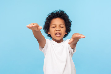 Portrait of blind little curly boy walking with closed eyes and outstretched hands, confused disoriented child with vision problems searching way. indoor studio shot isolated on blue background