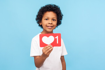 Wonderful charming little boy with curly hair holding one heart Like icon, button of social network to appreciate content, follower notification. indoor studio shot isolated on blue background