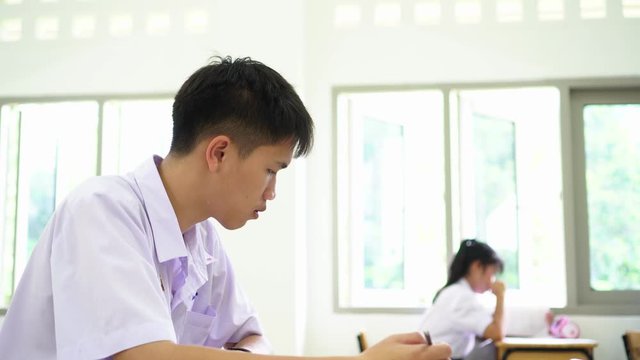 Student young man testing in exercise taking fill in exam paper answer sheet with students group at school or university in test room, document exams at campus classroom, education uniform of Thailand