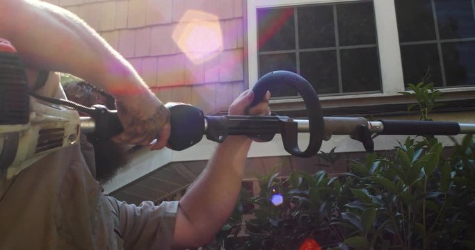 Landscape worker using a hedge trimmer to prune a holly bush beside a house