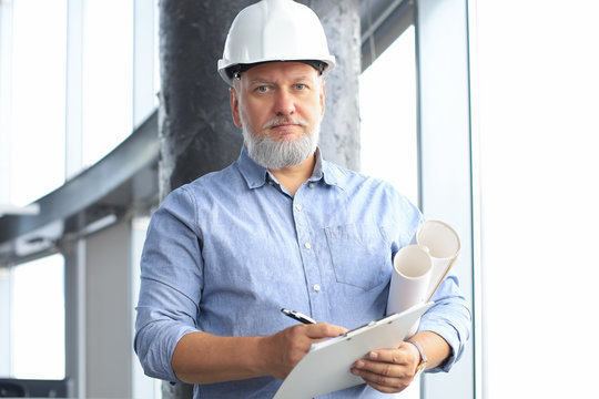 Confident mature business man in hardhat holding blueprint while standing indoors.