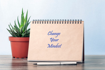 Motivational caption: "Change your mindset." On the table is a flower in a pot, there is an open notebook, near the notebook is a pen.