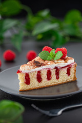 Piece of Poke Cake with raspberries on a black plate, mintleafs and green plants in the background