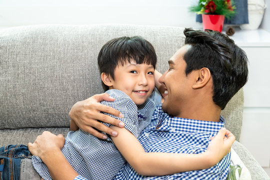 Happy Asian Father Hug And Cuddling His Son Lying On Sofa In Living Room, Family Bonding And Relationship Concept; Boy Looking At Camera