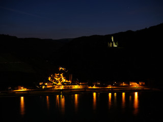Night shot of Castle Maus, colorfully illuminated, in front of the Rhine valley and under a starry sky. Below the small city of Wellmich