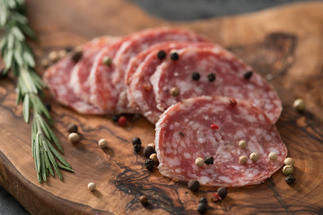 Sliced salame milano sausage on olive wood board with rosemary and pepper