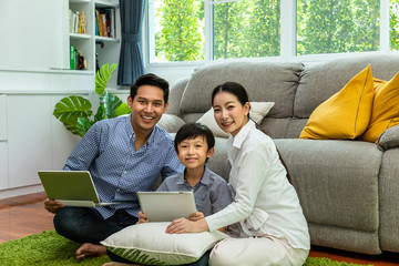 Happy Asian parent sitting together with boy on floor in living room; father working on laptop, and son using tablet with mother; looking at camera
