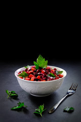 A bowl with Russian salad vinaigrette on a black background with copy space. Fork and sprigs of parsley nearby