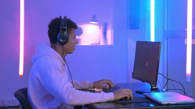 Profile view of young gammer in headphones playing in online shooter. The room have warm colored neon light.