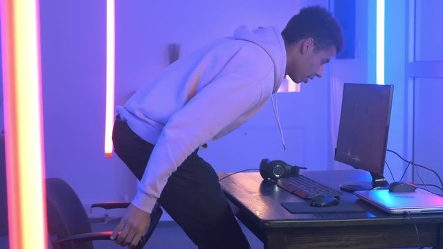 Black young gamer sitting to the table, taking earphones on and starting playing online shooter. He playing game on his personal computer.