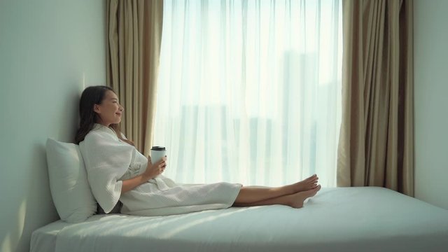 A young Asian woman dressed in a robe relaxes on the bed while preparing to drink from a disposable coffee cup.