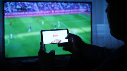 Background of the TV at home (at the bar), the guy orders different snacks, pizza and sweets scrolling through internet stores using mobile phones. Concept: Sport TV.