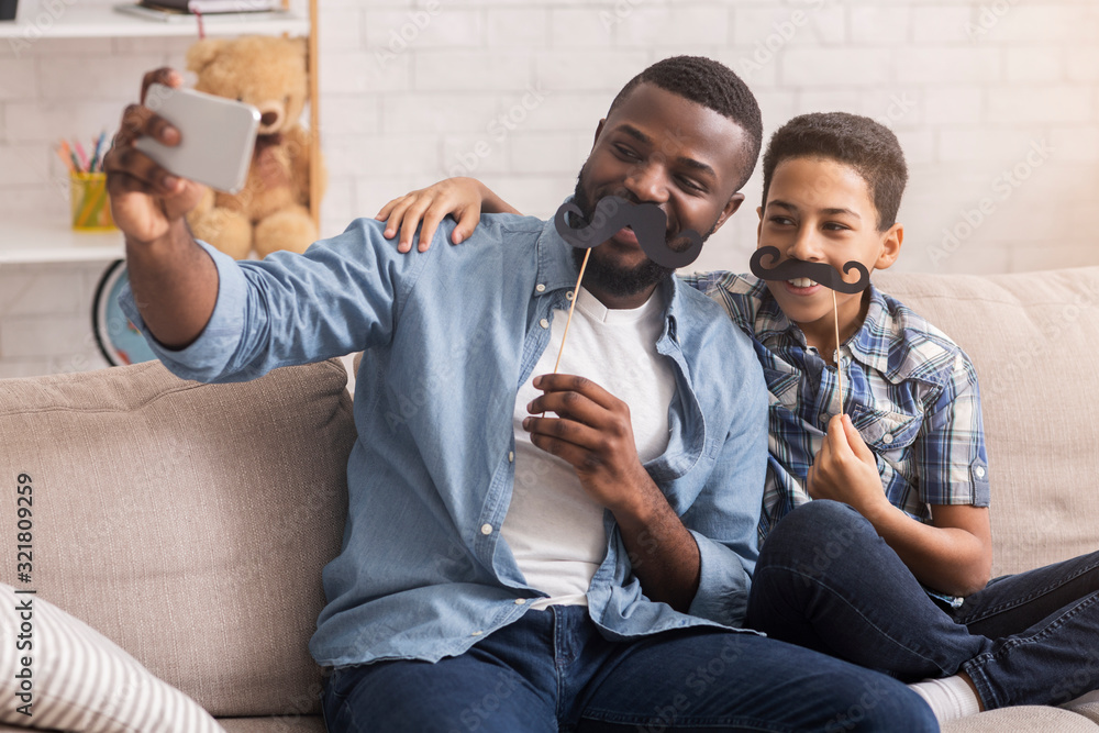 Wall mural Joyful black father and son posing for selfie at home