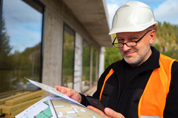 A construction worker in a white helmet smiles at the camera. A man is holding construction drawings and a mobile phone. The civil engineer is going to call to the customer of works.