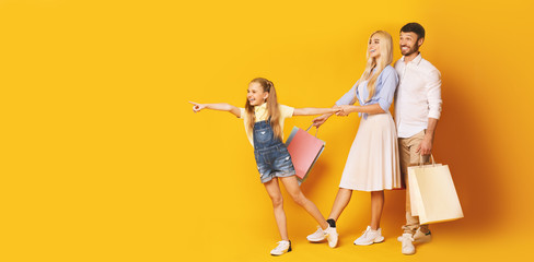 Little Daughter Pointing Finger Aside Shopping With Parents, Yellow Background