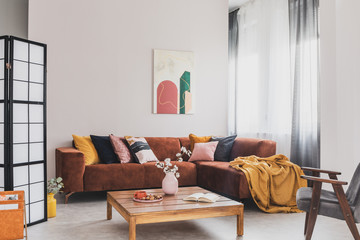 Real photo of a yellow blanket lying on a brown corner sofa in bright living room interior with a wooden table and a gray chair