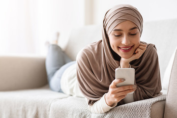 Arab girl using smartphone while lying on cozy couch at home