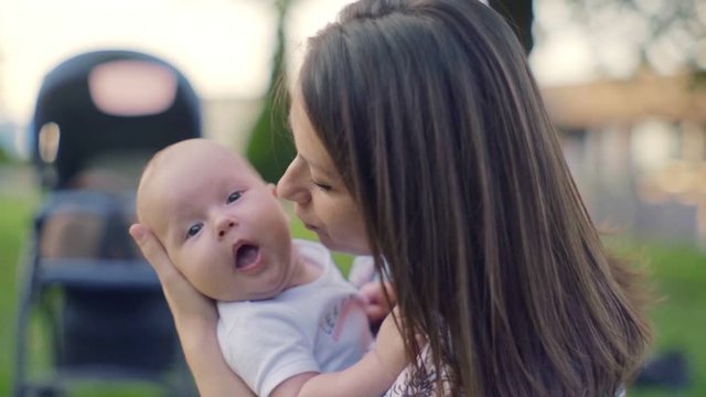 Mother with a child outdoors in a park. Young brunette woman holding  three month old baby boy in her arms and kisses him. In the background is a stroller. Slow motion, 100 fps.