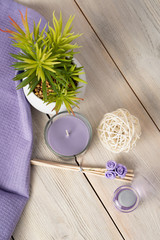 Spa composition of a bottle of aromatic oil, candles, a flower in a pot, a straw ball, wooden incense sticks with roses and a purple napkin on a wooden background.