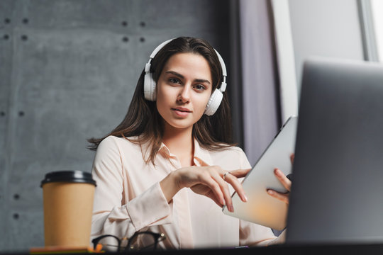 Young woman sitting at desktop in wireless headphones and listening to music, webinar, language lesson. Digital tools and gadgets for work in office. Young girl enjoying entertainment content online.