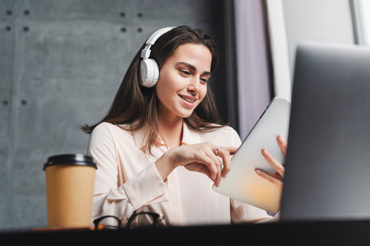 Millennial young woman enjoying great sound of music in wireless headphones. Great visual and audio content for channel subscribers. Girl typing on laptop and texting on smart tablet.
