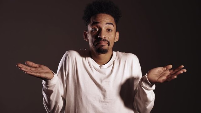 Portrait of unsure african man in white shrugs his arms, makes gesture of I don't know, care. Body language