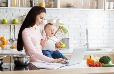 Young mom typing on laptop, sitting with baby at kitchen