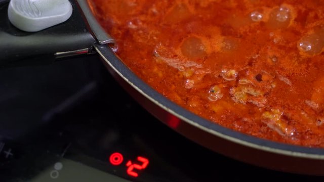 Red liquid is cooked in black frying pan on electric stove. bolognese dressing is being prepared on fire.