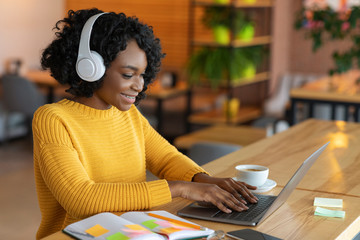 Happy black girl in headphones looking at laptop, cafe interior