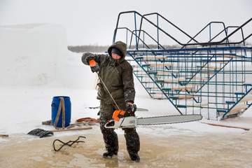 Worker in a green jacket with a hood with a chainsaw in his hands