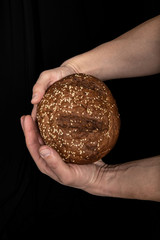 Man's hands hold tasty fresh loaf of dark bread with sesame seeds on black background